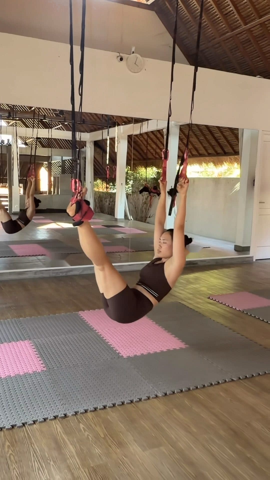 A woman hangs on the lianas in the Canggu studio, the loops holding her weight, a mirror and a thatched roof behind her