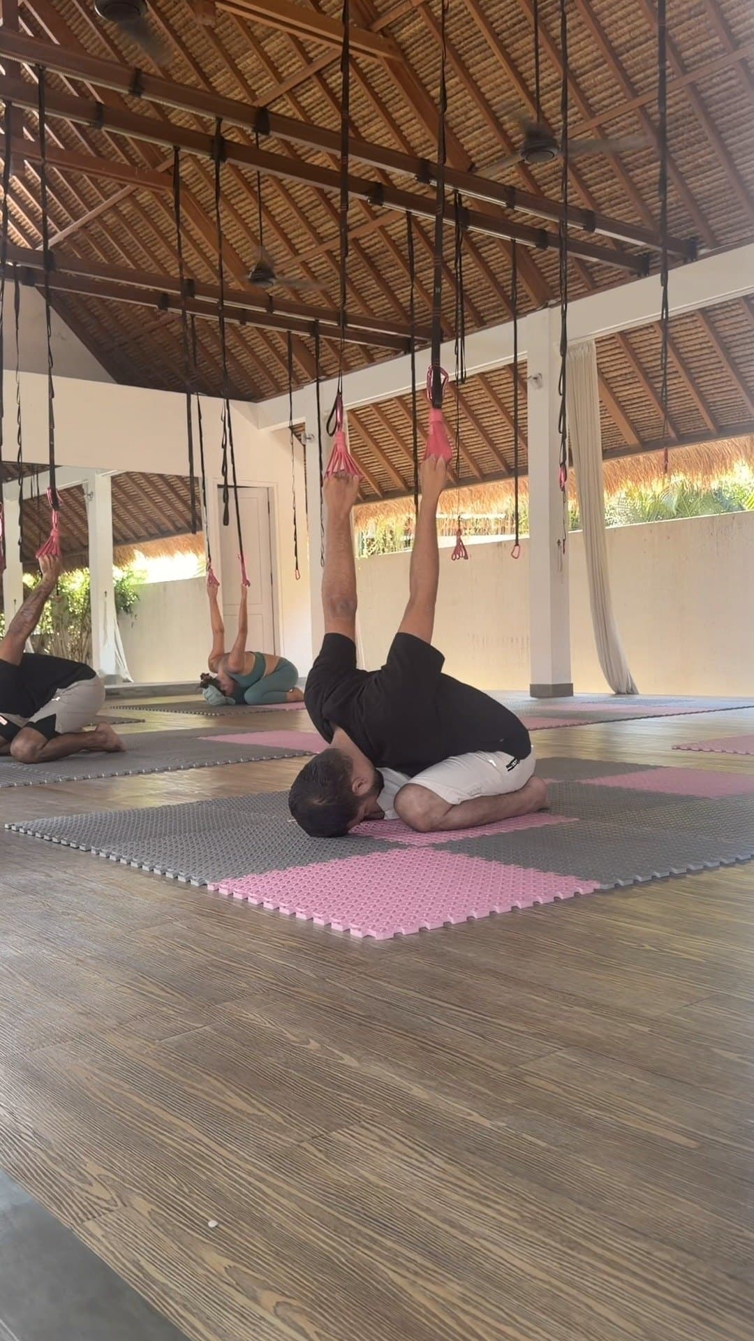 A group class: several people hang on the lianas under a thatched roof, a man among them